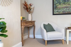 Ein Zimmer mit einem weißen Sessel, einem Holznachttisch mit einer Vase getrockneter Blumen und einem gerahmten Bild an der Wand.