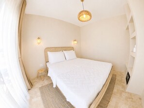 Een slaapkamer met een groot bed, twee wandlampen, een hanglamp, een klein nachtkastje en een geweven vloerkleed.