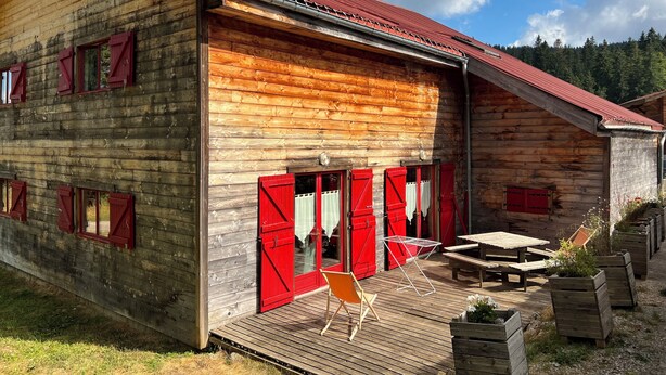 A wooden building with red shutters, a wooden deck, and a metal table. - Chalet in the Les Rousses resort, between Prémanon and LAMOURA (PREMANON)