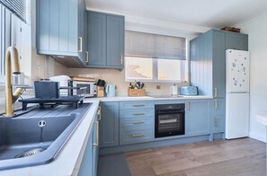 A modern kitchen with grey cabinets, a built-in oven, a sink, and a window with blinds. - Points, an art deco Wittering family seaside holiday home for 10 guests (East Wittering)