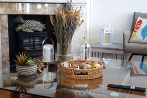A living room with a fireplace, a glass coffee table, a potted plant, a woven basket, and a remote control. - Points, an art deco Wittering family seaside holiday home for 10 guests (East Wittering)