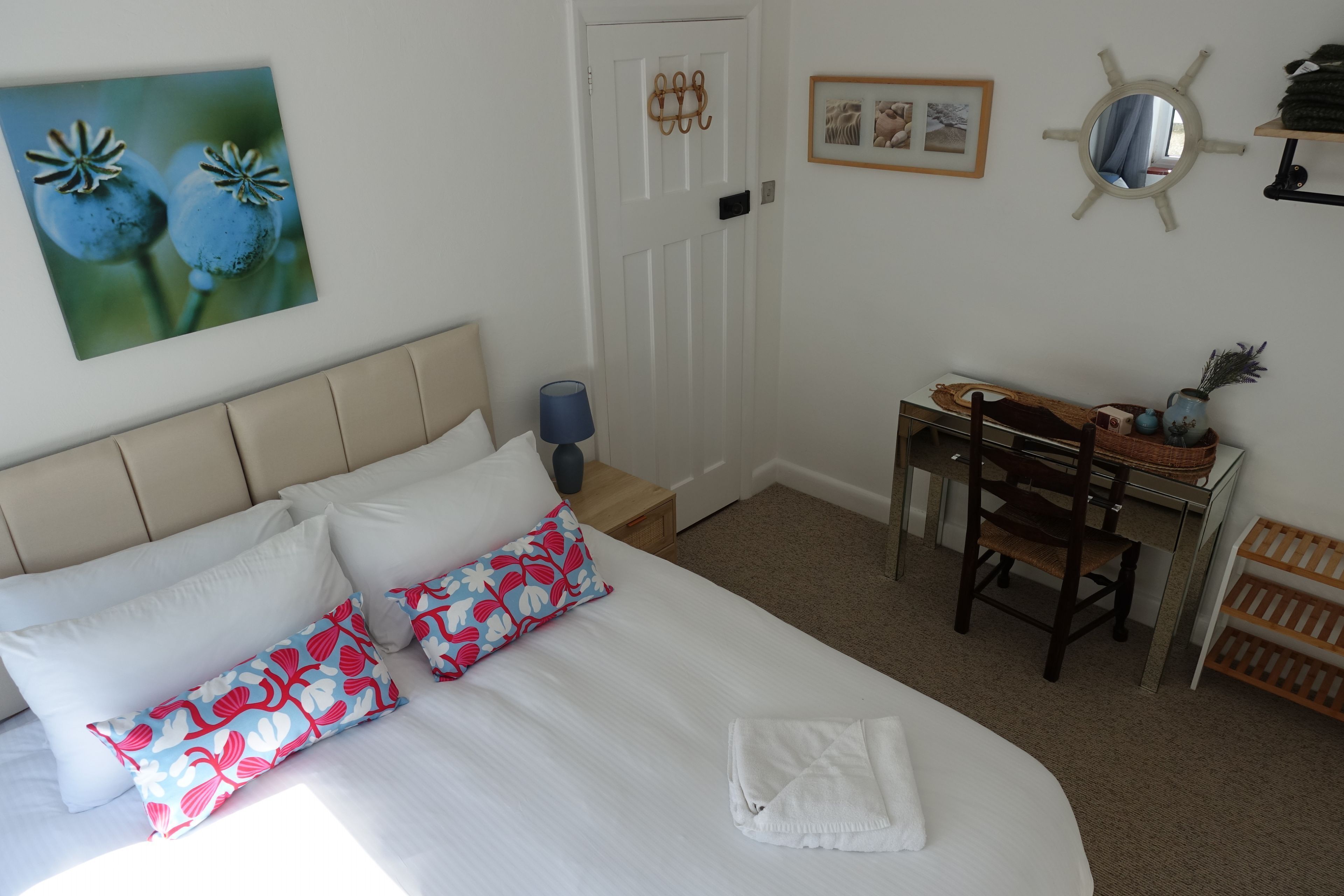A neatly made bed with white and floral pillows, a wooden chair, a desk with a vase, and a framed picture on the wall.