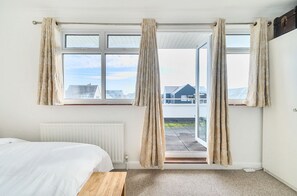 A bedroom with a bed, a radiator, a window with curtains, and a view of buildings outside. - Points, an art deco Wittering family seaside holiday home for 10 guests (East Wittering)