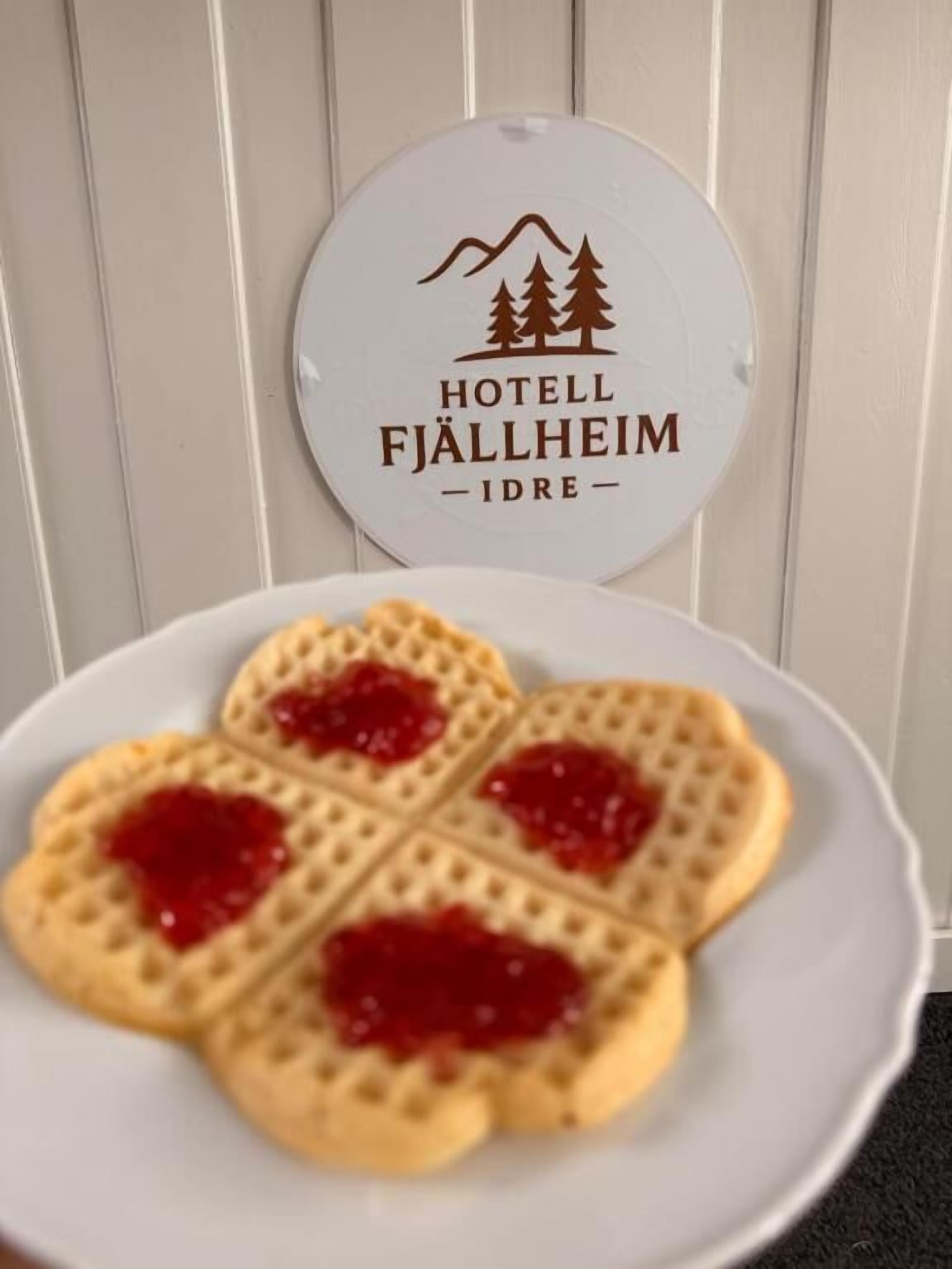 Ein Teller herzförmige Waffeln mit roter Marmelade, ein Schild für Hotel Fjällheim Idre und ein weißer Holzhintergrund.
