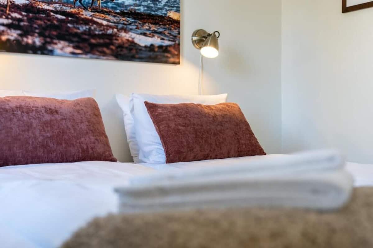 A neatly made bed with white and brown pillows, a wall-mounted lamp, and a painting of a snowy landscape.