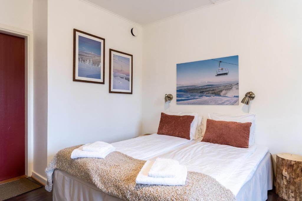 Una cama doble con sábanas blancas, dos cuadros en la pared y un ventanal con vistas a un paisaje nevado y un telesilla.
