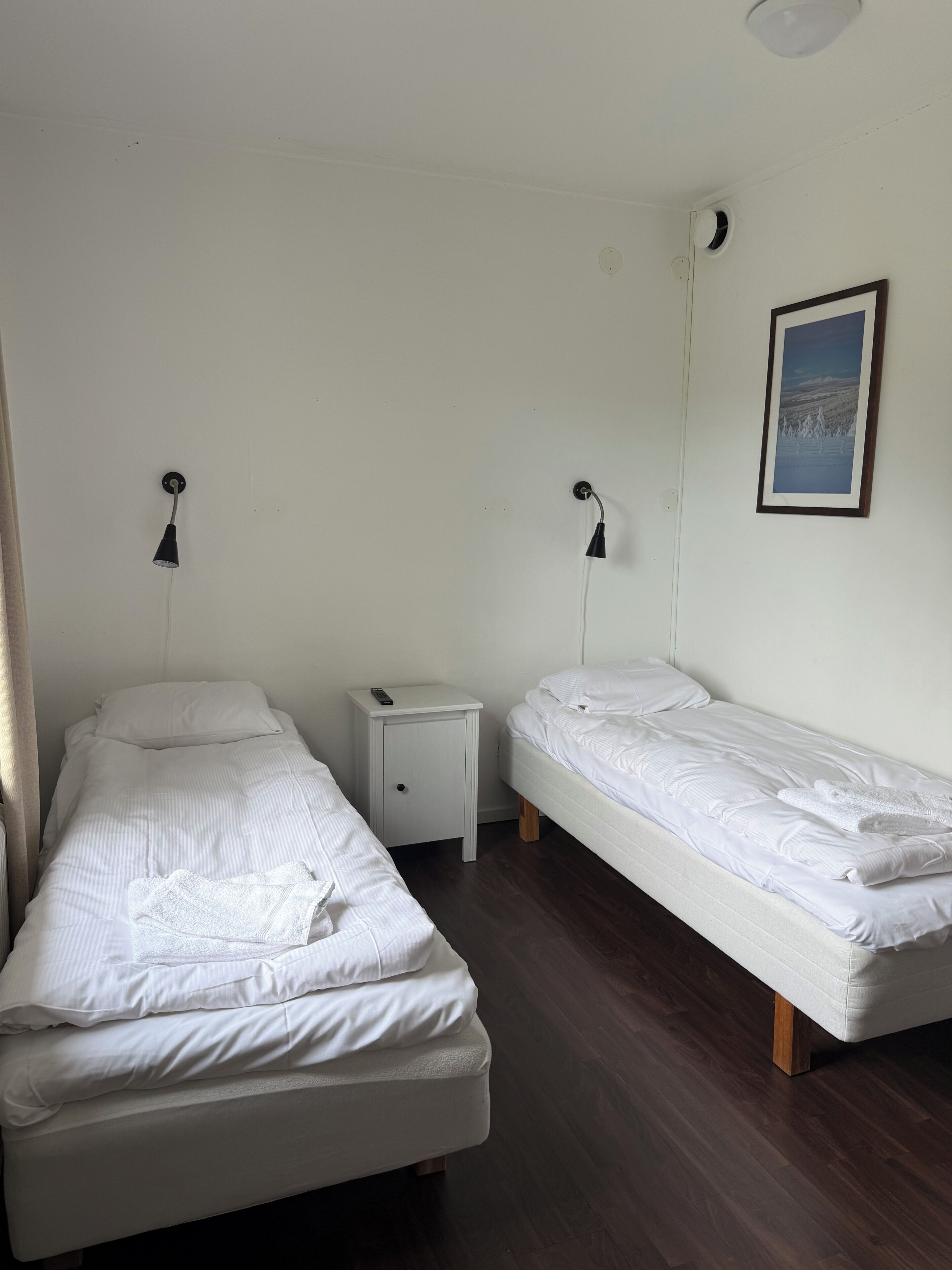 A room with two single beds, a small bedside table with a remote control, a wall-mounted light, and a framed picture.