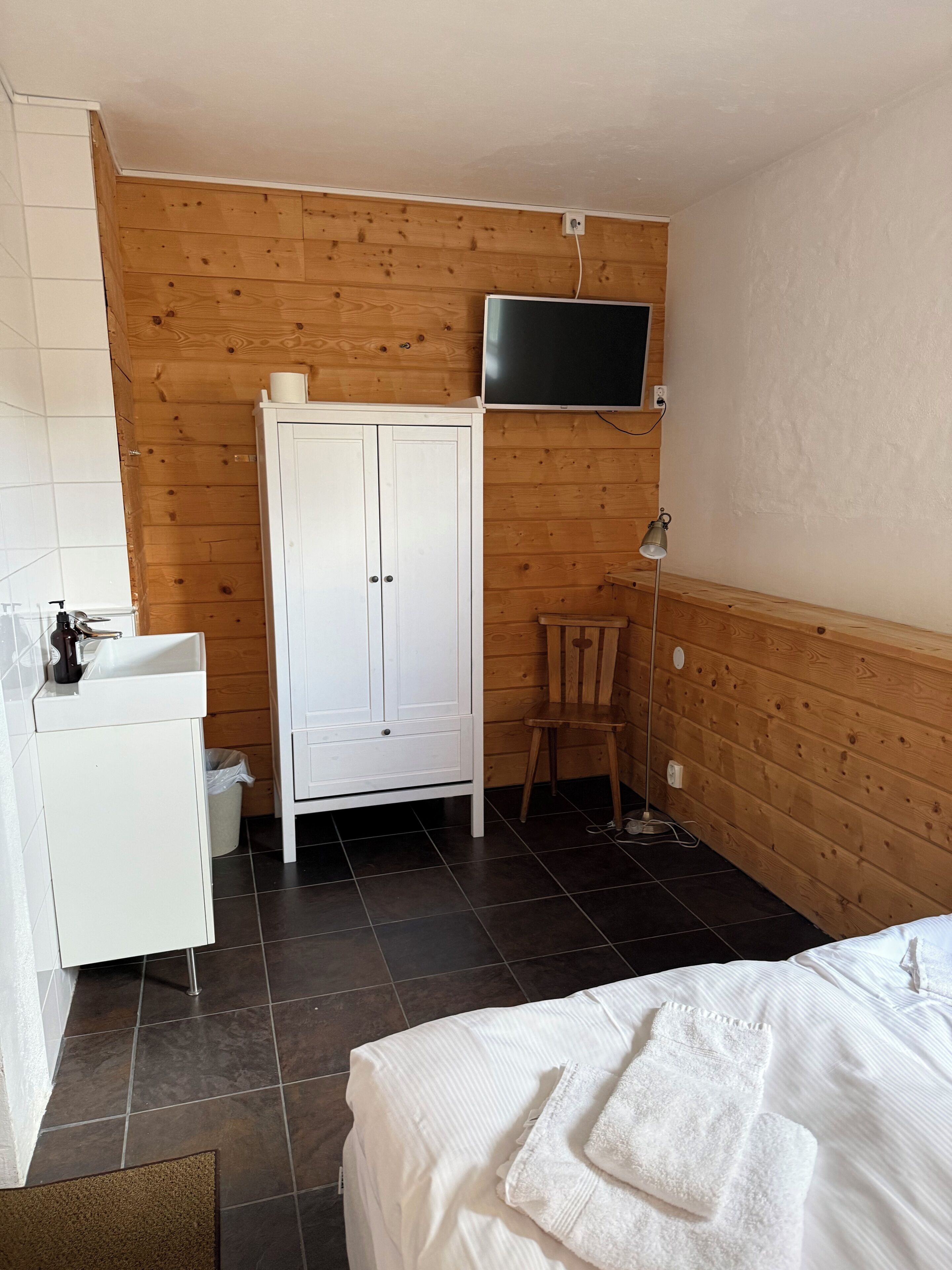 A room with a bed, a white wardrobe, a wooden cabinet, a television mounted on the wall, and a chair.