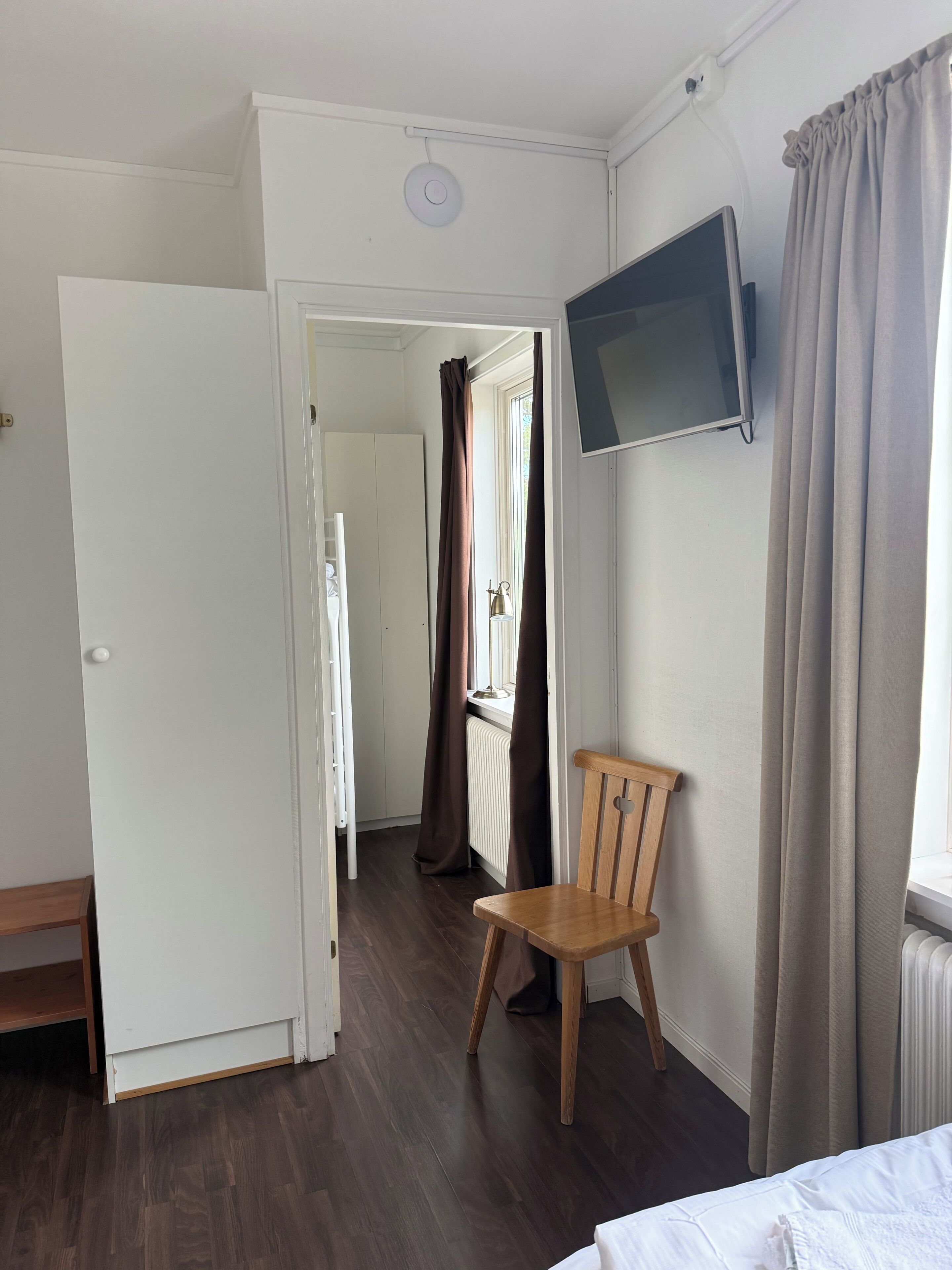 Una habitación con una cama, un televisor montado en la pared, una silla, piso de madera y una puerta que da a otra habitación.