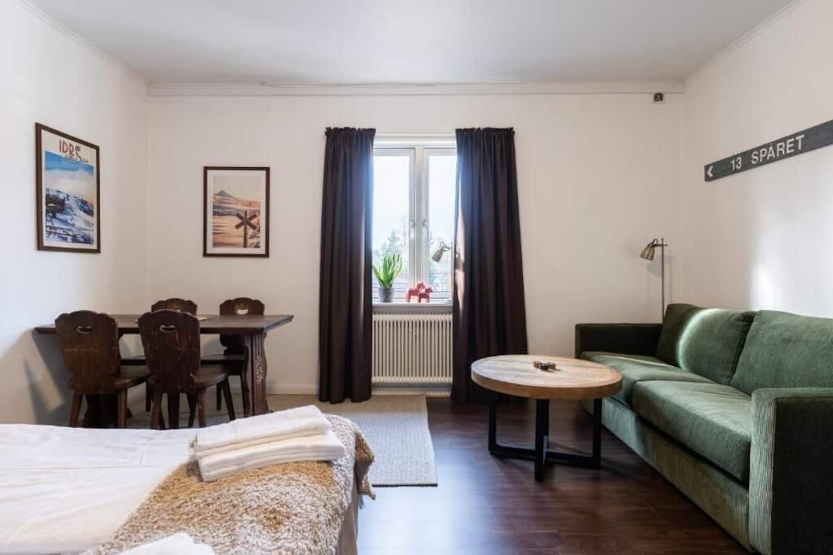 A hotel room with a bed, a green sofa, a round table, a dining table with chairs, a radiator, and a window with curtains.