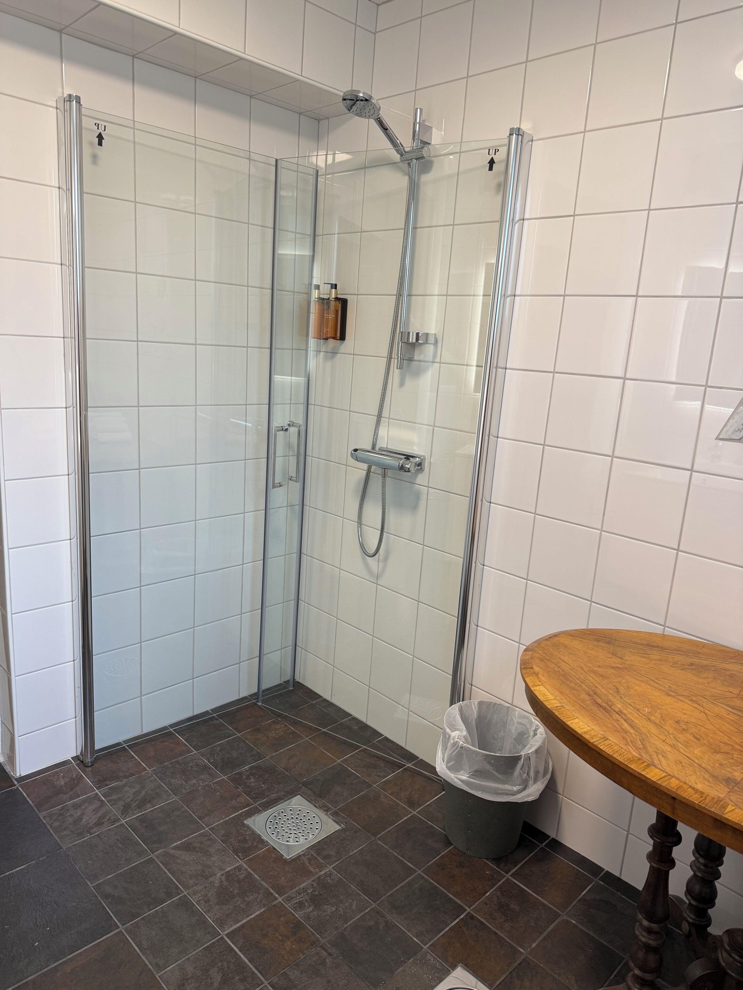 Eine Dusche mit Glastür, ein Holztisch und ein Papierkorb.