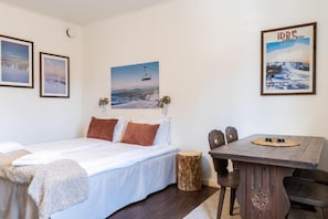 Un dormitorio con una cama, una mesa de madera, sillas, un taburete pequeño y cuadros en la pared.