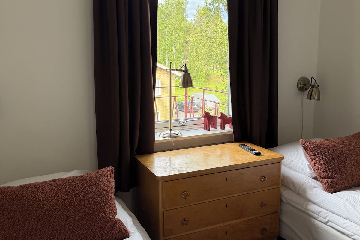A room with two beds, a wooden dresser, a window with curtains, a lamp, and a view of the outside.