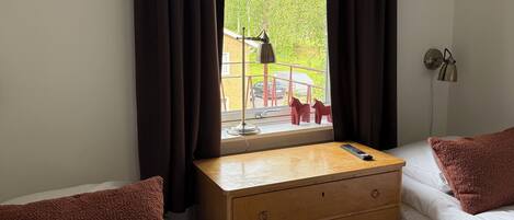 A room with two beds, a wooden dresser, a window with curtains, a lamp, and a view of the outside.