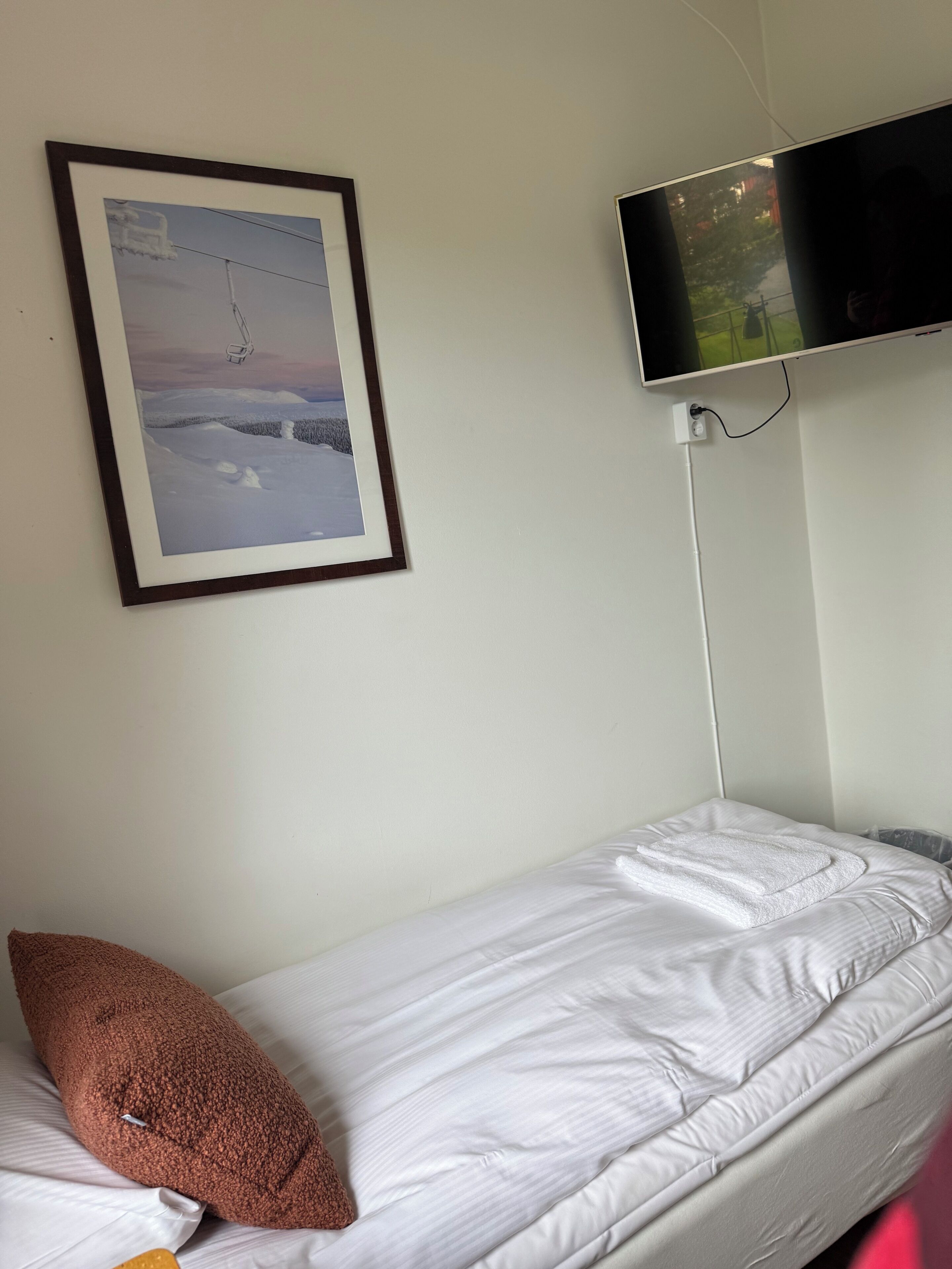 A single bed with white bedding, a brown pillow, a framed picture of a snowy landscape, and a mounted television on the wall.