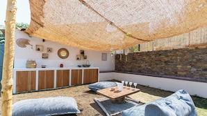 Une terrasse extérieure avec une table en bois, deux fauteuils en forme de sac de couchage de couleur grise, un auvent en fibres tressées et un mur en pierre.