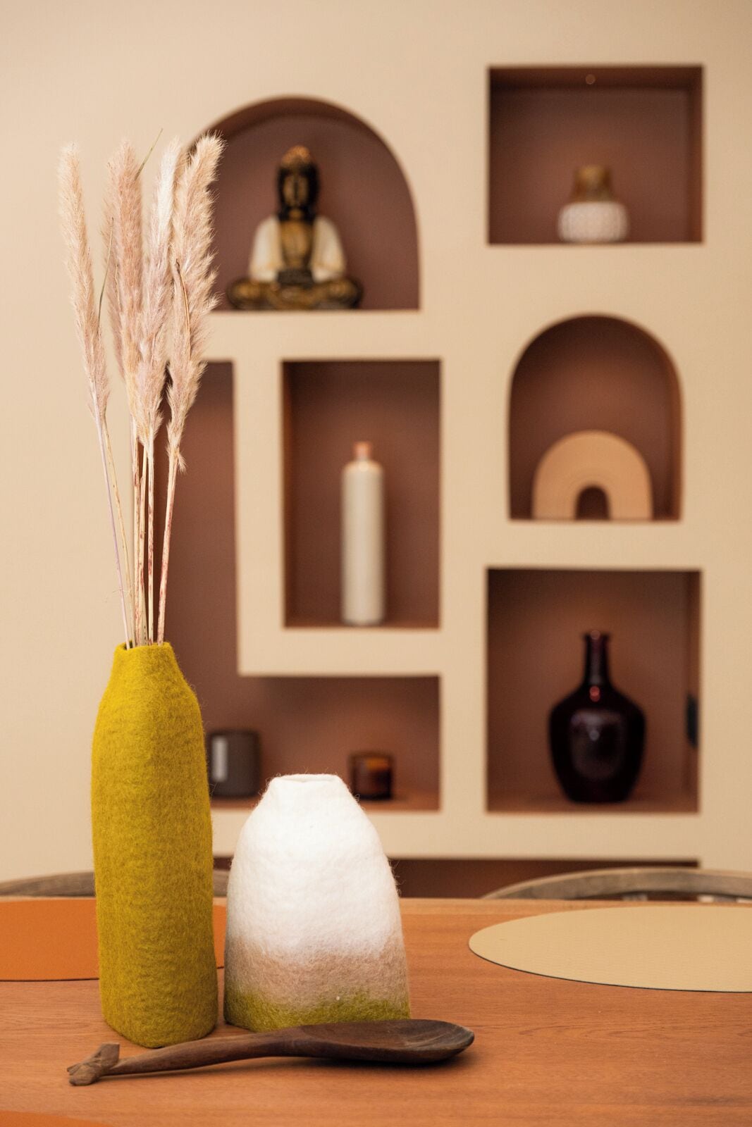 Une pièce avec une table en bois, un vase vert recouvert de feutre contenant des plumes d’herbe des pampas et une sculpture blanche également recouverte de feutre. Des étagères en arrière-plan présentent divers objets décoratifs.