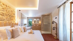 Une chambre spacieuse avec un grand lit, un mur en pierre, une salle de bain avec douche et une vue sur l’extérieur.