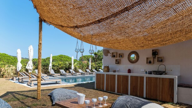Un espace aménagé au bord de la piscine, avec des chaises longues, une table en bois sur laquelle sont posés des verres à vin, et un coin bar doté de meubles en bois.