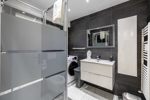 Ein modernes Badezimmer mit Duschkabine aus Glas, integrierter Waschmaschine, Waschtisch mit Spiegel und Handtuchhalter.