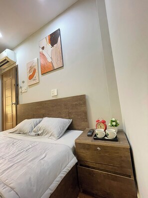 Un dormitorio con cabecera de madera, cama con ropa de cama blanca, mesita de noche con un reloj y una planta pequeña, y una obra de arte en la pared.