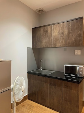 Una cocina compacta con armarios de madera, una encimera negra, un microondas integrado, un ventilador blanco y un refrigerador.