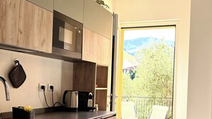 Une cuisine moderne équipée d’un four encastré, d’une bouilloire et d’une machine à café. Une fenêtre donne sur un balcon offrant une vue sur la verdure.