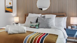 Een net opgemaakt bed met kussens, een gestreepte wand, een ronde spiegel, een ingelijste foto en een nachtkastje met een lamp.