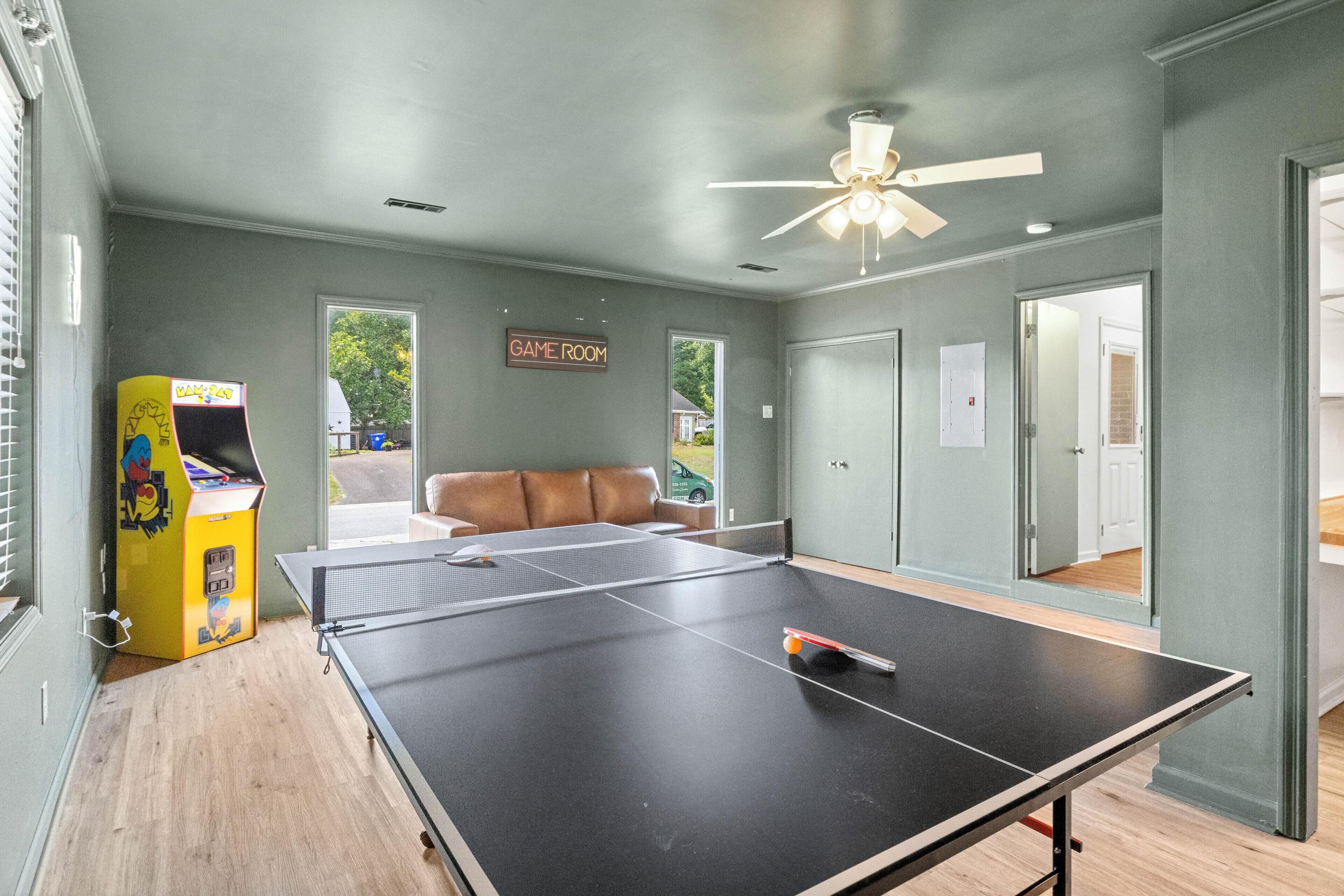 Una habitación con mesa de ping pong, sofá, ventilador de techo y puerta que da a otra habitación.