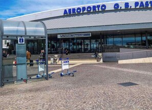 Una terminal de aeropuerto con un cartel que dice 'AEROPORTO G, B, PASTO' y un área de espera techada con bancos y un mostrador de venta de boletos.