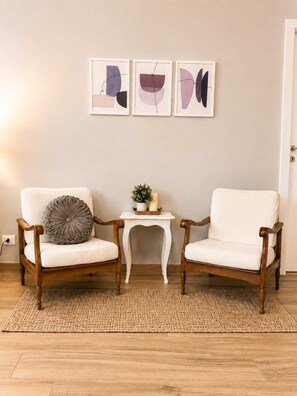 Ein Zimmer mit zwei weißen Sesseln, einem kleinen Tisch mit einer Pflanze und drei abstrakten Wandbildern.