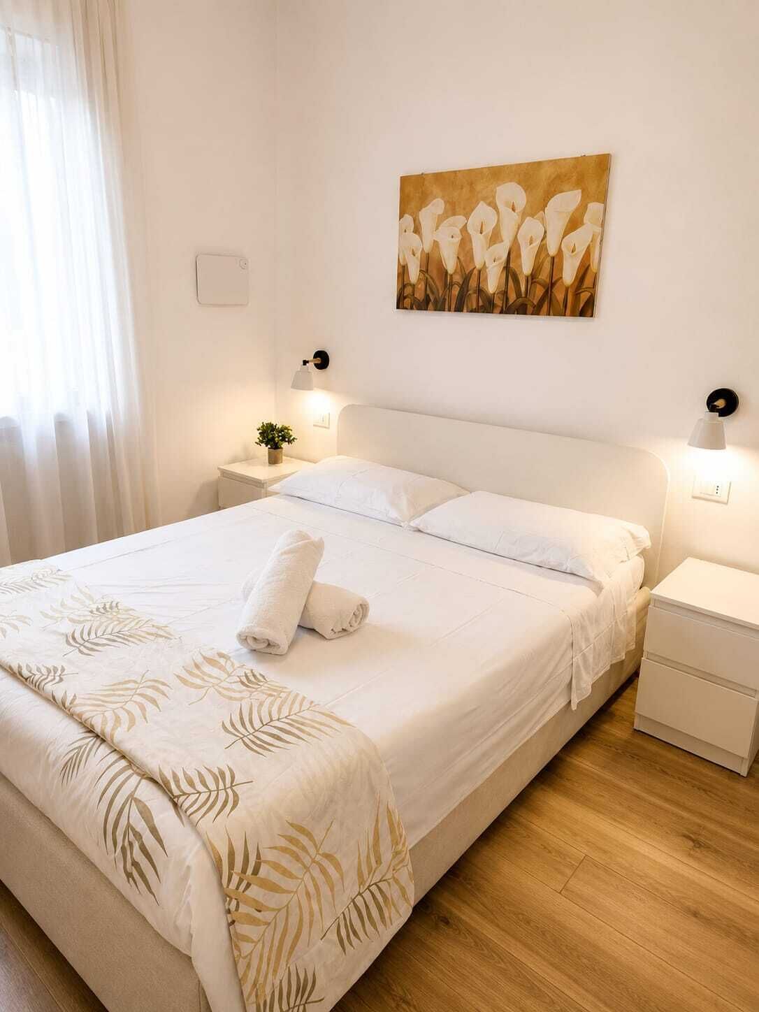 A neatly made bed with a white comforter and a rolled towel, a bedside table with a lamp, and a wall-mounted light fixture.