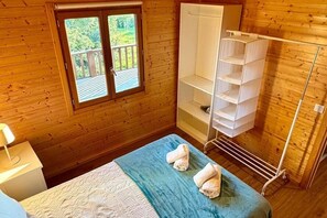 A wooden-paneled room with a bed, a window with a view of greenery, a white wardrobe, and a drying rack.
