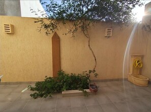 Une cour avec un sol carrelé, un petit arbre, un jardinière et un tuyau d’arrosage jaune.