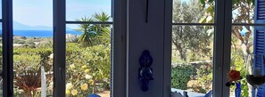 A room with a view of a garden and the sea, a wall-mounted light fixture, and a vase with a flower. - Beautiful apartment in the middle of the flowering garden with sea view (Naxos)