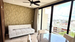 Een moderne woonkamer met een bank, een ronde eettafel en stoelen. Er is een plafondventilator, een groot raam met uitzicht op de stad, en een behang met een patroon.