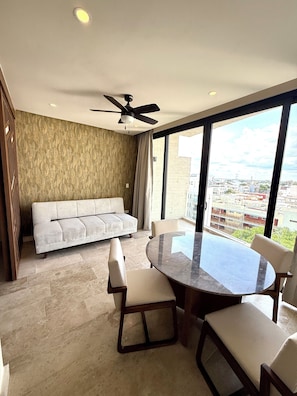 Ein modernes Wohnzimmer mit einer Couch, einem runden Esstisch und Stühlen. Ein Deckenventilator, ein großes Fenster mit Stadtblick und eine Wand mit Musterung sind ebenfalls vorhanden.
