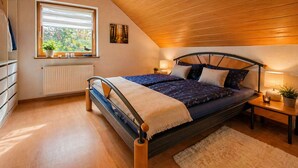 A bedroom with a sloped ceiling, a bed with a headboard, a window with blinds, a radiator, and a bedside table with a lamp. - Spacious vacation apartment in a quiet location with lots of nature. Pet friendly! (Tiefenthal)
