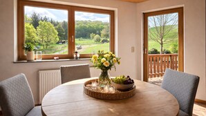 A room with a round table, two chairs, a vase with flowers, a bowl of fruit, and a view of greenery through large windows. - Spacious vacation apartment in a quiet location with lots of nature. Pet friendly! (Tiefenthal)