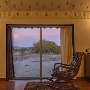 A room with a sliding glass door leading to a desert landscape, a rocking chair, and a small table with a kettle. - Innara Hospitality (Bali)