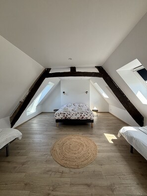 A bedroom with a sloped ceiling, a bed with a floral bedspread, a woven rug, and a small table with a lamp. - Gîte La Carelle in Vains at the foot of the Bay of Mont Saint Michel (Vains)