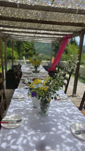 A long table set with plates, cutlery, and floral centerpieces under a pergola with a view of greenery.