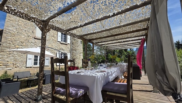 Un espace extérieur aménagé pour les repas, avec une pergola couverte, un bâtiment en pierre et des tables dressées.