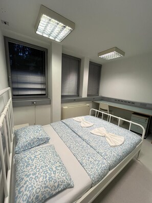 Quarto com cama, mesas de cabeceira, escrivaninha e janela com persianas.