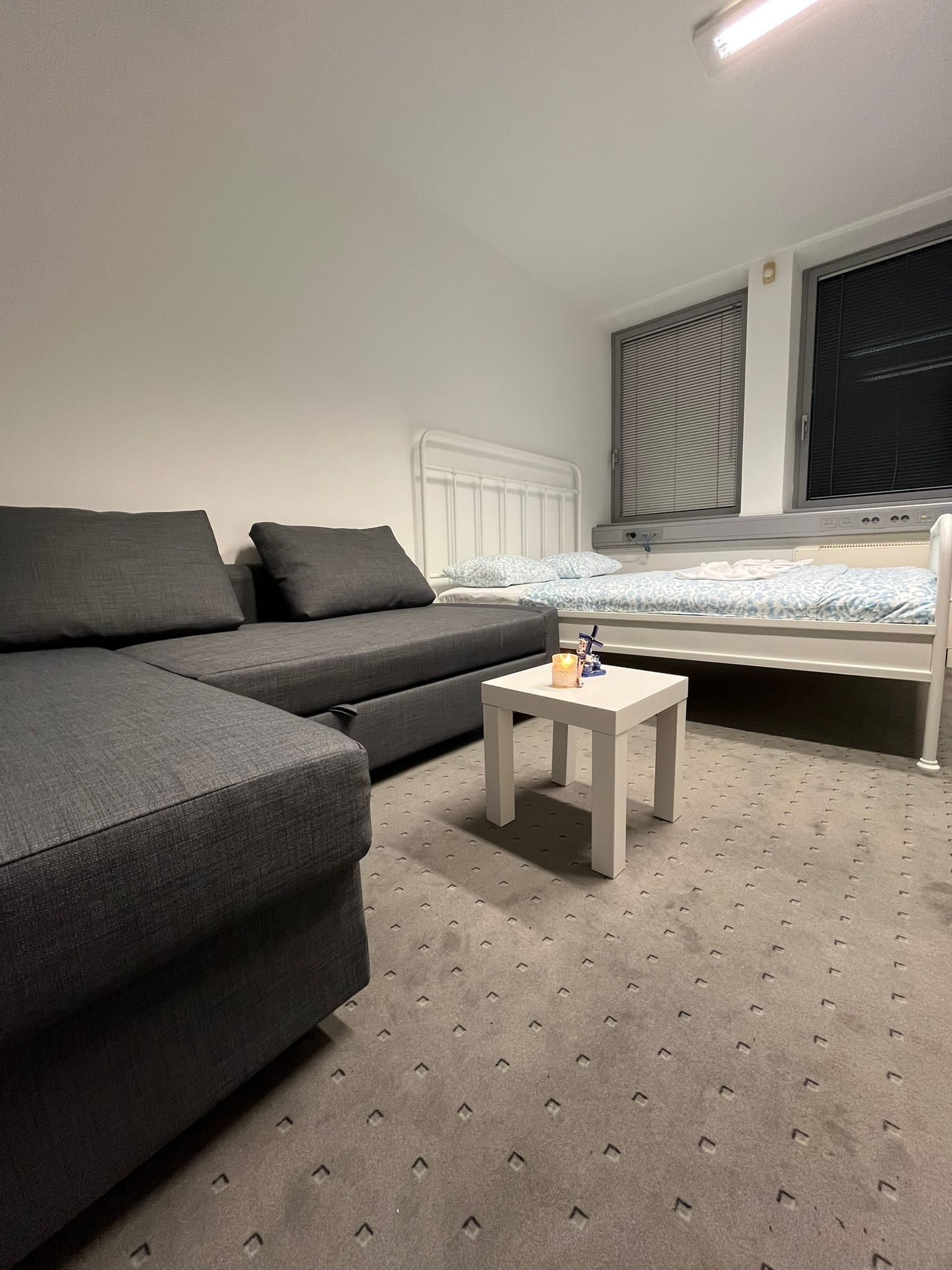 Une chambre moderne avec un canapé gris, une petite table blanche, un lit avec une couette à motifs et une fenêtre avec des stores.
