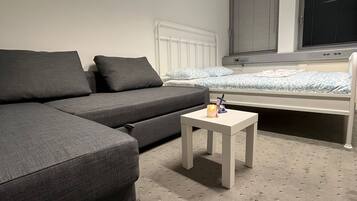 Une chambre moderne avec un canapé gris, une petite table blanche, un lit avec une couette à motifs et une fenêtre avec des stores.