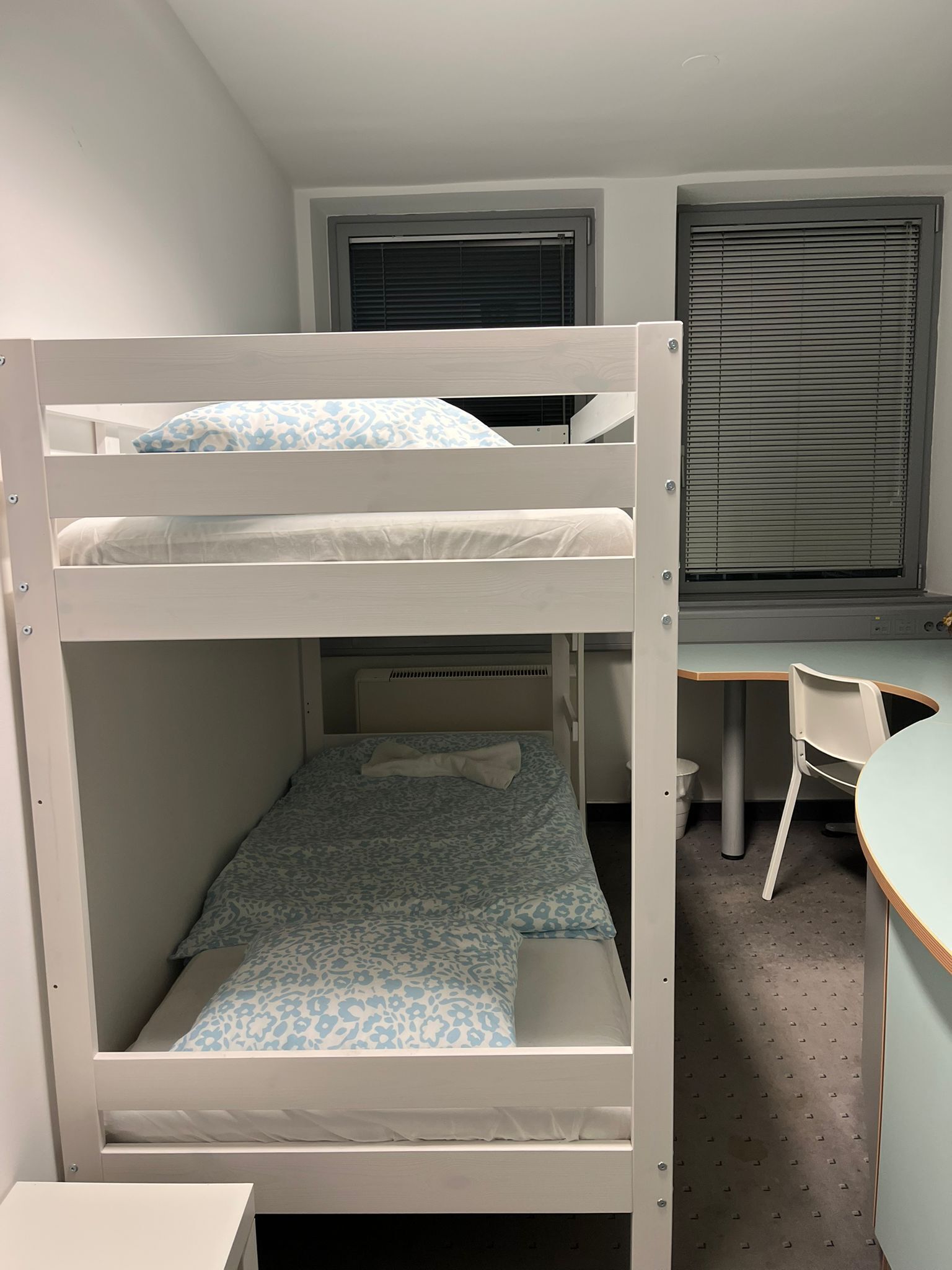 A bunk bed with a desk and chair, a window with blinds, and a radiator.