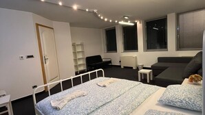 Ett modernt hotellrum med en enkel säng, ett litet sängbord, en stol, en soffa och ett fönster med persienner.