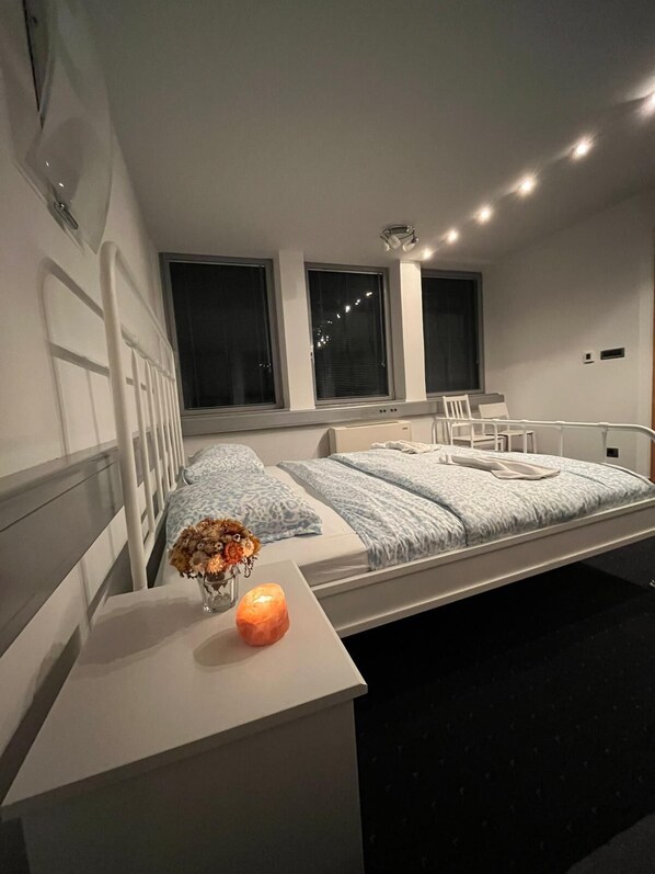 Quarto moderno com cama, mesa de cabeceira e um criado-mudo com um vaso e vela.