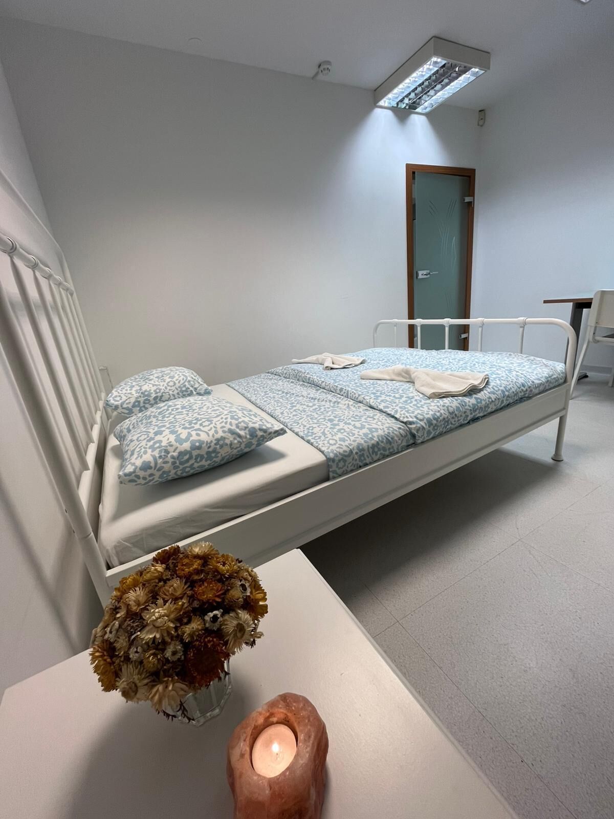 A hospital room with a single bed, a small table with a candle and flowers, and a door in the background.